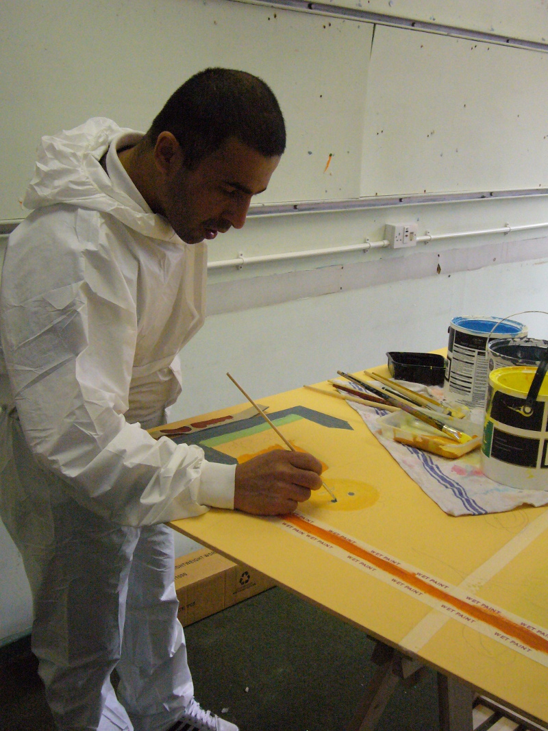 Ahmad is painting his mural design onto boards inside a room. He has short dark hair and is wearing a white jump-suit with the hood down.