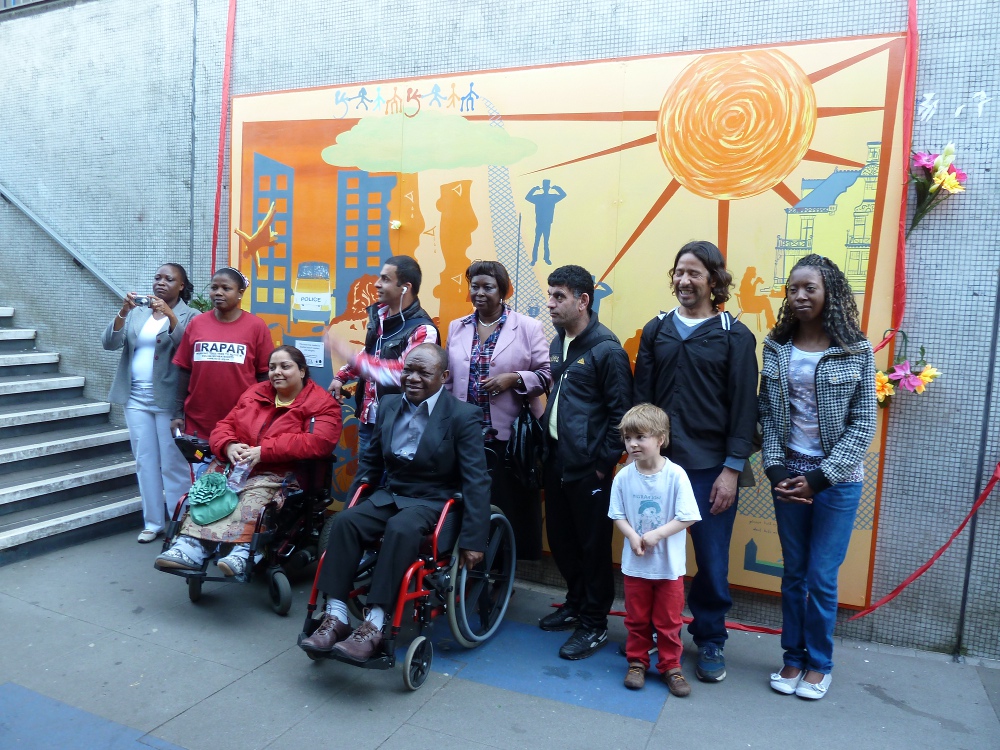 This picture shows all the participants standing in front of the mural. There are nine adults, five women and four men, and one little boy. Two of the participants, Manjeet and David, are wheelchair users.
