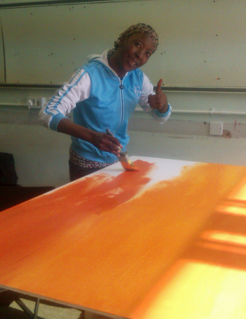 Lucie is looking at the camera and smiling, giving a thumbs-up. She is inside a room and is wearing a blue and white tracksuit top and a headscarf. She is painting the board's background of oranges and yellows.