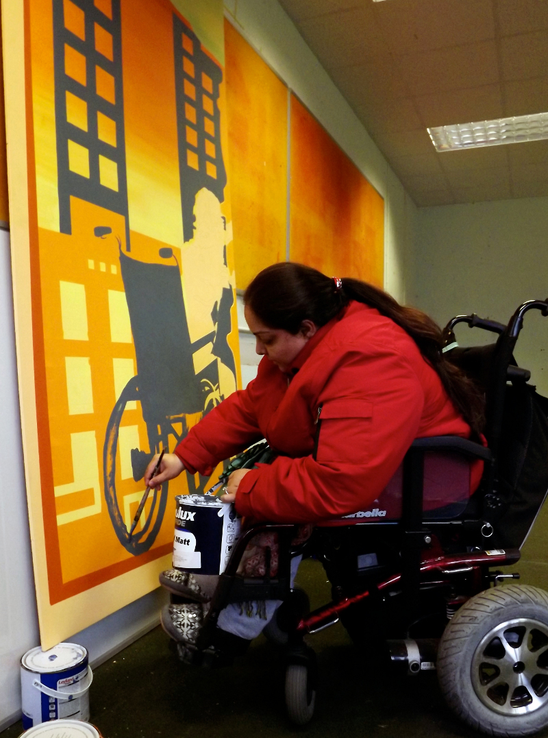 Manjeet is inside painting the mural design onto the boards. Manjeet is a wheelchair user, she is holding a can of paint, painting a blue wheelchair onto an orange background. Manjeet has long dark hair in a pony-tail and she is wearing a red coat.
