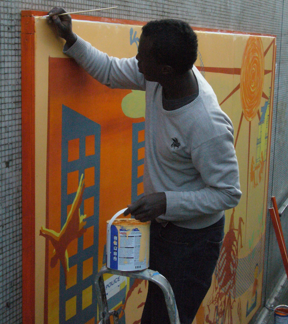 A man is painting the orange border of the mural now that it has been installed in the subway. He is standing on a step-ladder and is holding a paint can. He has short dark hair and is wearing a beige jumper.
