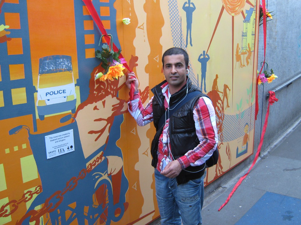 This picture is of Ahmad cutting the ribbon. The ribbon is red and has bunches of yellow and purple flowers along it. Ahmad has is wearing white headphones and is wearing a red-checked shirt and blue jeans with a black body warmer over the shirt.