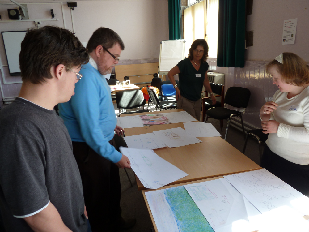 Four people are standing round a table, two men and two women. The man at the front of the pictures has a grey t shirt, short hair and is wearing glasses. Behind him is a bearded man with a light blue jumper on.  He is also wearing glasses. At the far end of the table a woman is walking round. She has wavy dark hair and is wearing a black t shirt. At the other side of the table there is a young woman wearing a cream coloured hairband and matching long sleeved jumper. They are all looking at pieces of paper on the table with drawings on them. The drawings are not really visible.