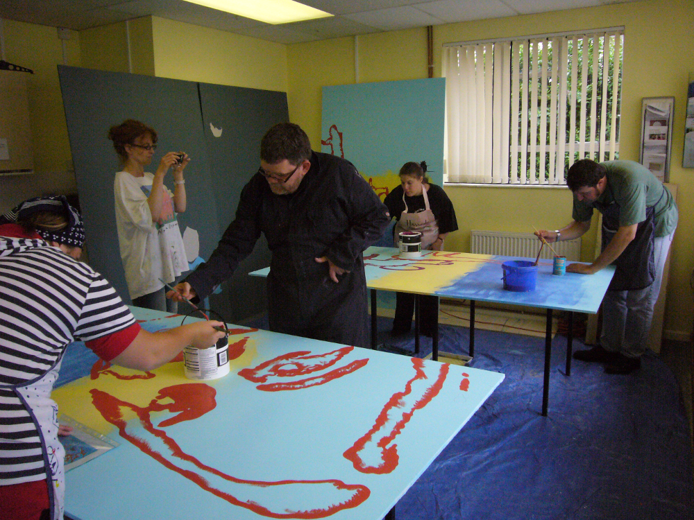 In this picture four people are busy painting on boards laid across tables. The peoples' faces are not clearly visible as they are bent over the boards. There is a man and a woman working on the board at the front, and a man and a woman working at the back. Another woman is standing with a camera. The board at the front of the picture is painted with a pale blue background and a large red silhouette of a face. The board at the back is painted in 3 stripes ? dark blue at the bottom, yellow in the middle and pale blue at the top.