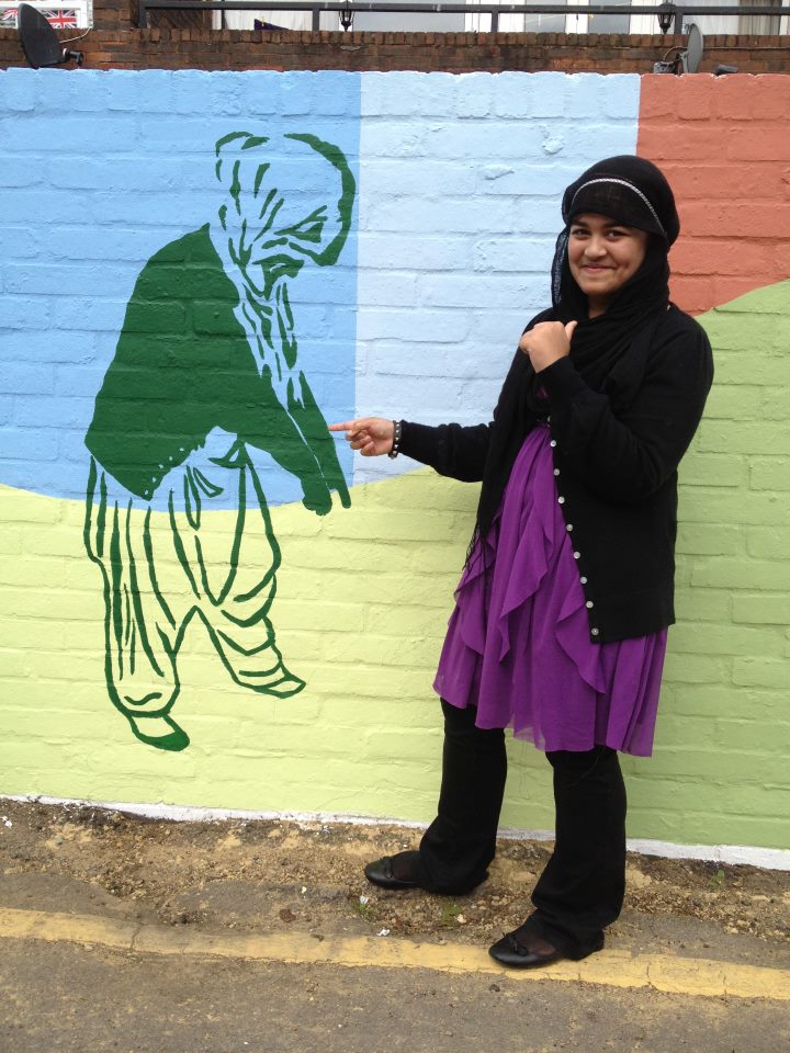 The photo is of a teenage girl who is looking at the camera whilst pointing to a figure that is part of the mural. The figure is herself, painted in green on a background of blue and yellow pointing at the ground.