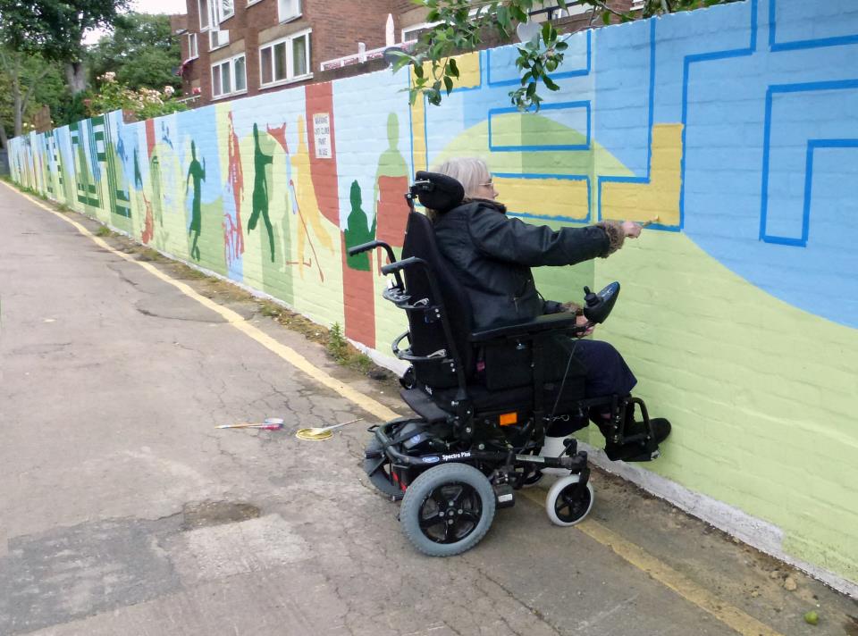 In this picture there is a woman with glasses and mid length grey hair painting the mural. The woman is a wheelchair user and is painting the letter S in yellow.