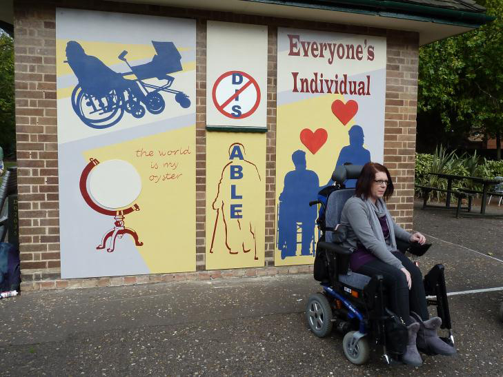 This photograph shows a woman sitting in a wheelchair in front of the painted mural. The mural is in three panels. On the right of the mural are the words 'Everyone's individual'. Below this are dark blue silhouettes of a wheelchair user holding hands with someone standing up. Red hearts are above their heads. The middle panel has the letters 'DIS' crossed out and beneath it the word 'ABLE' written vertically down the outline of a person using crutches. On the left there is a blue silhouette of a wheelchair user pushing a pram. Underneath this is a picture of a globe with the words 'The world is my oyster'.