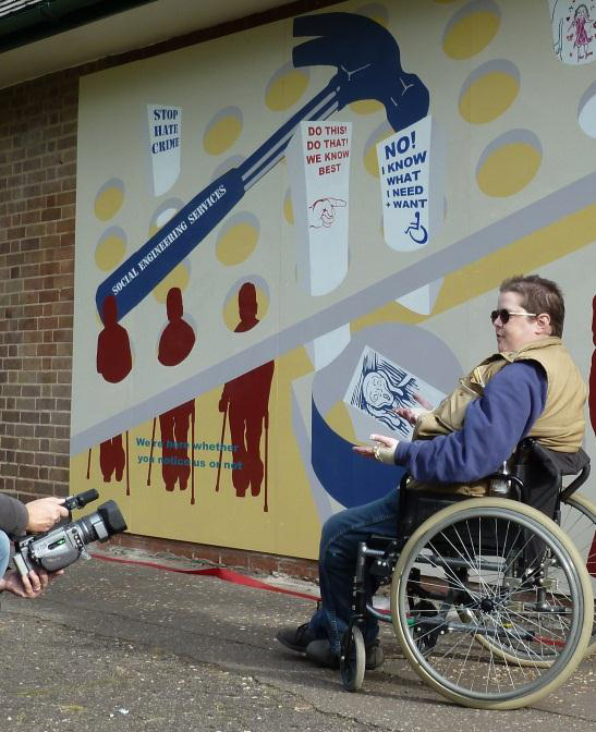 This picture shows a wheelchair user on the right hand side. She has short hair, is wearing glasses, a green sleeveless jacket on top of a blue jumper and jeans. She has her hands palms up in front of her and is talking in front of the section of the mural with the blue hammer and the square pegs being squashed through round holes. The image of the person screaming is in the bin just above her hands. 
A film camera can be seen at the bottom right.